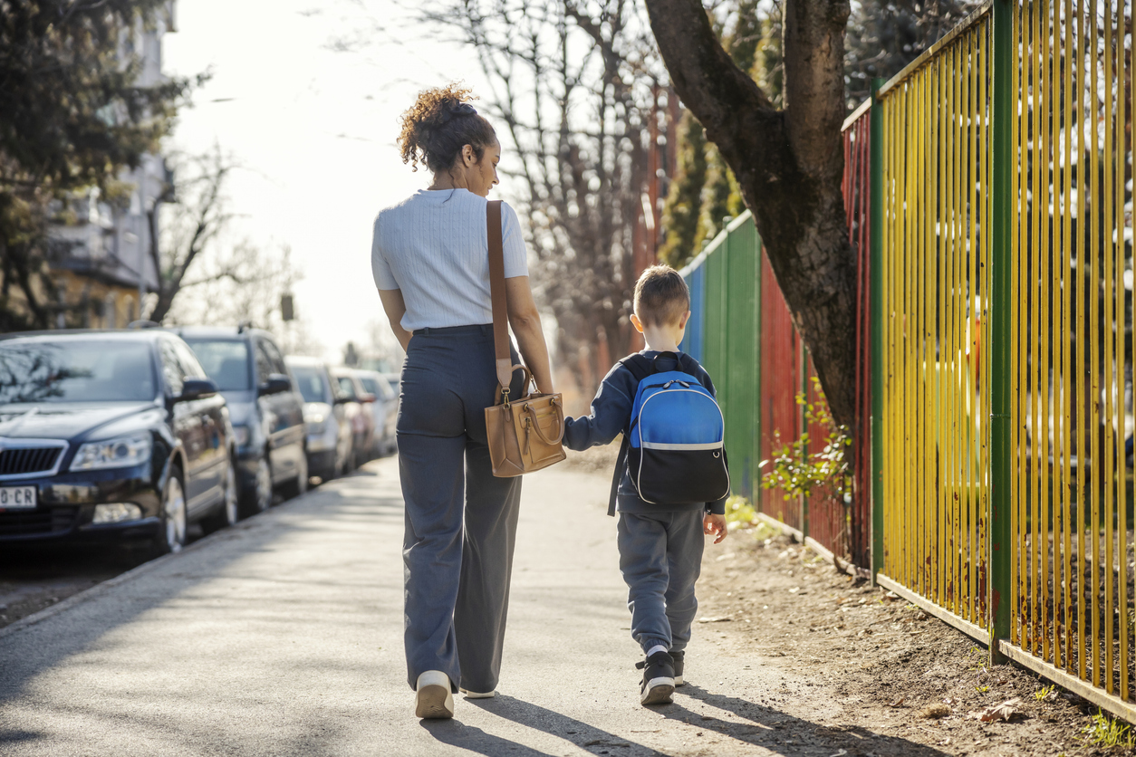 Rear view of young mother holding hands with her son and taking him to kindergarten.