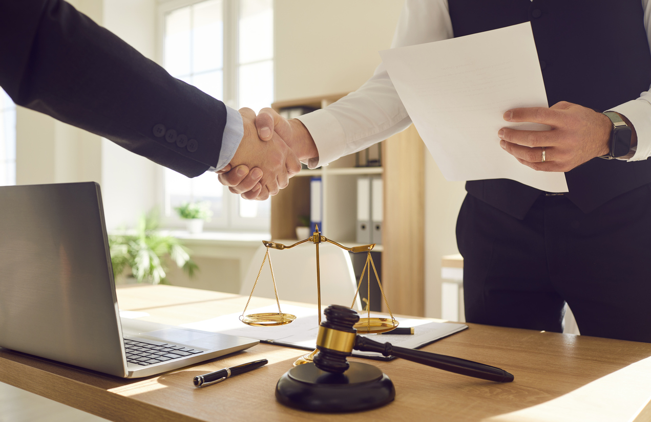 Cropped photo of a male lawyer working in office shaking hand with a man client standing at the desk on workplace with a laptop, judge's gavel and scales on table. Legal law and advice concept.