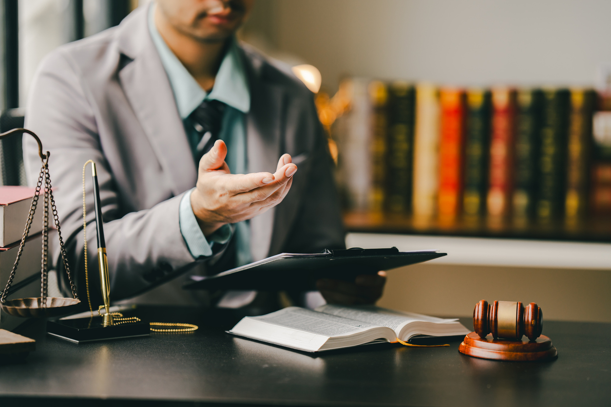 Legal professional in a suit discussing documents with an open hand gesture, sitting at a desk with an open law book, justice scales, and a gavel, representing legal consultation and advocacy