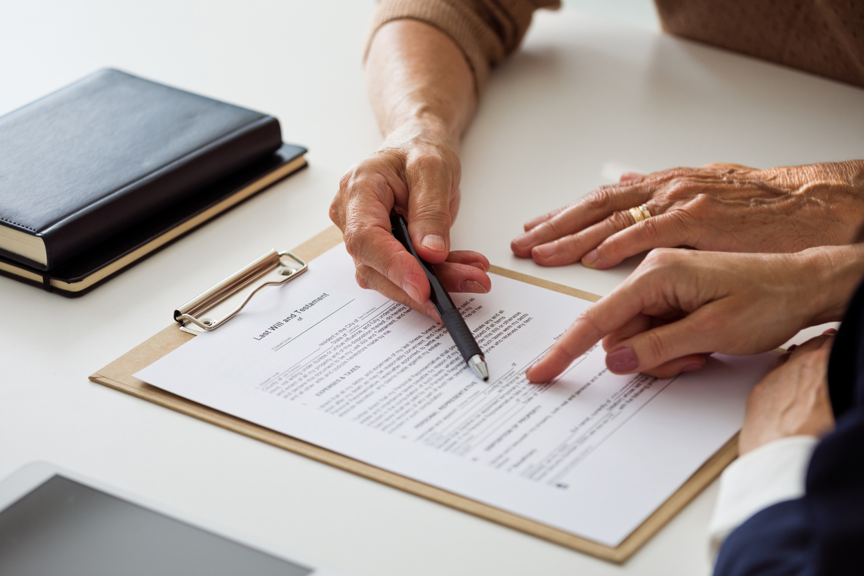 Professional lawyer pointing to legal document on clipboard while discussing last will and testament details with a senior client during a consultation.